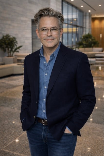 A person with glasses wearing a denim shirt and jacket.