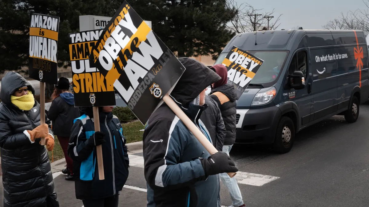 Amazon warehouse workers carry signs in a picket line in front of an Amazon supply chain facility. The signs read: "Amazon is Unfair" and "Amazon: Obey the Law."