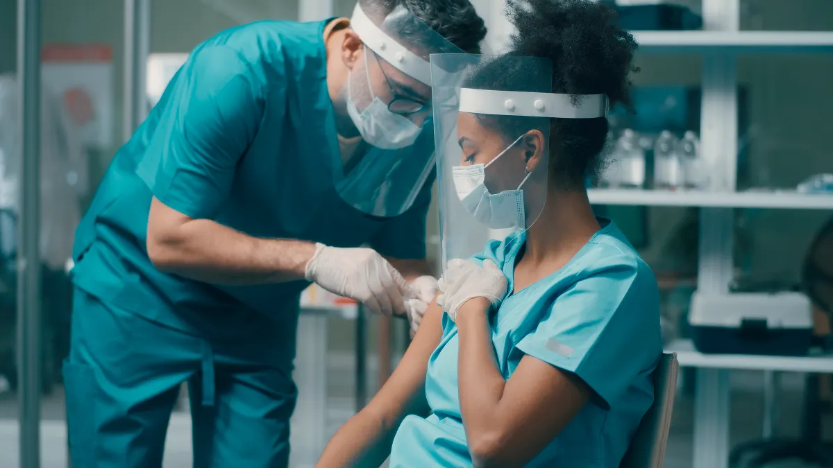 Male medical practitioner making vaccine injection to ethnic female colleague while working in hospital together
