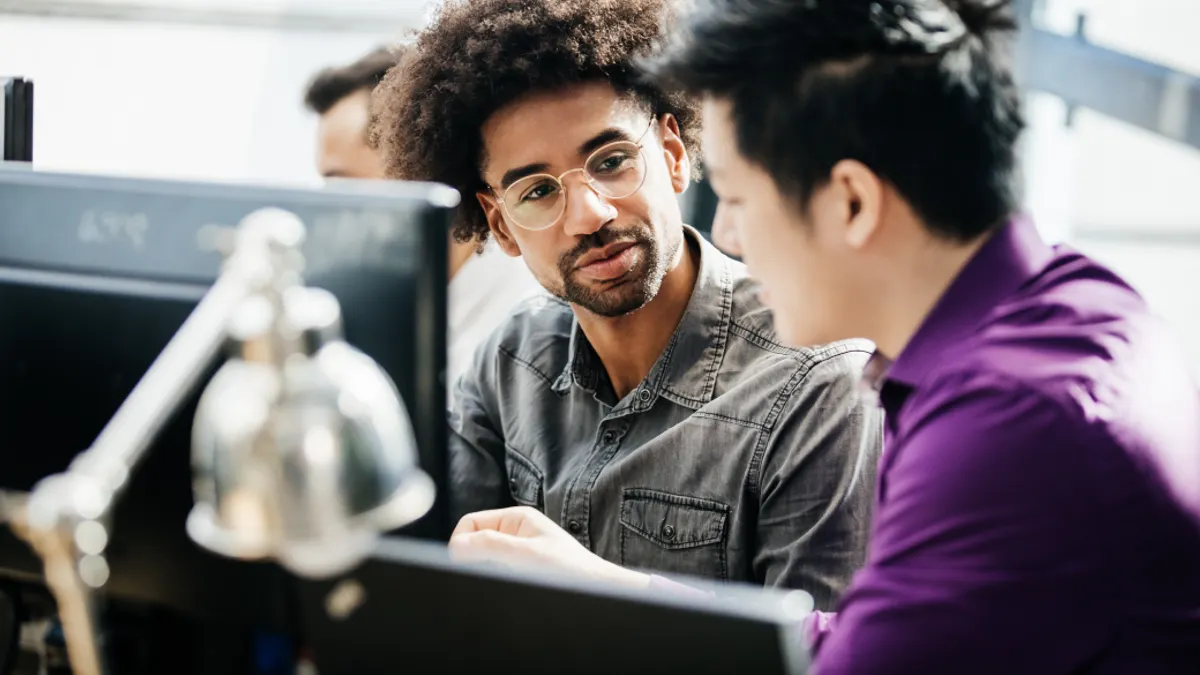 Two men are engaged in discussion at a workstation. One wears glasses and a denim shirt, the other a purple shirt. A lamp and computer monitors are visible.
