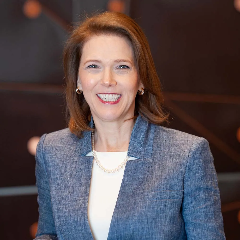 An image of a woman with auburn hair smiling. She is wearing a gray suit, a white dress shirt and pearls.