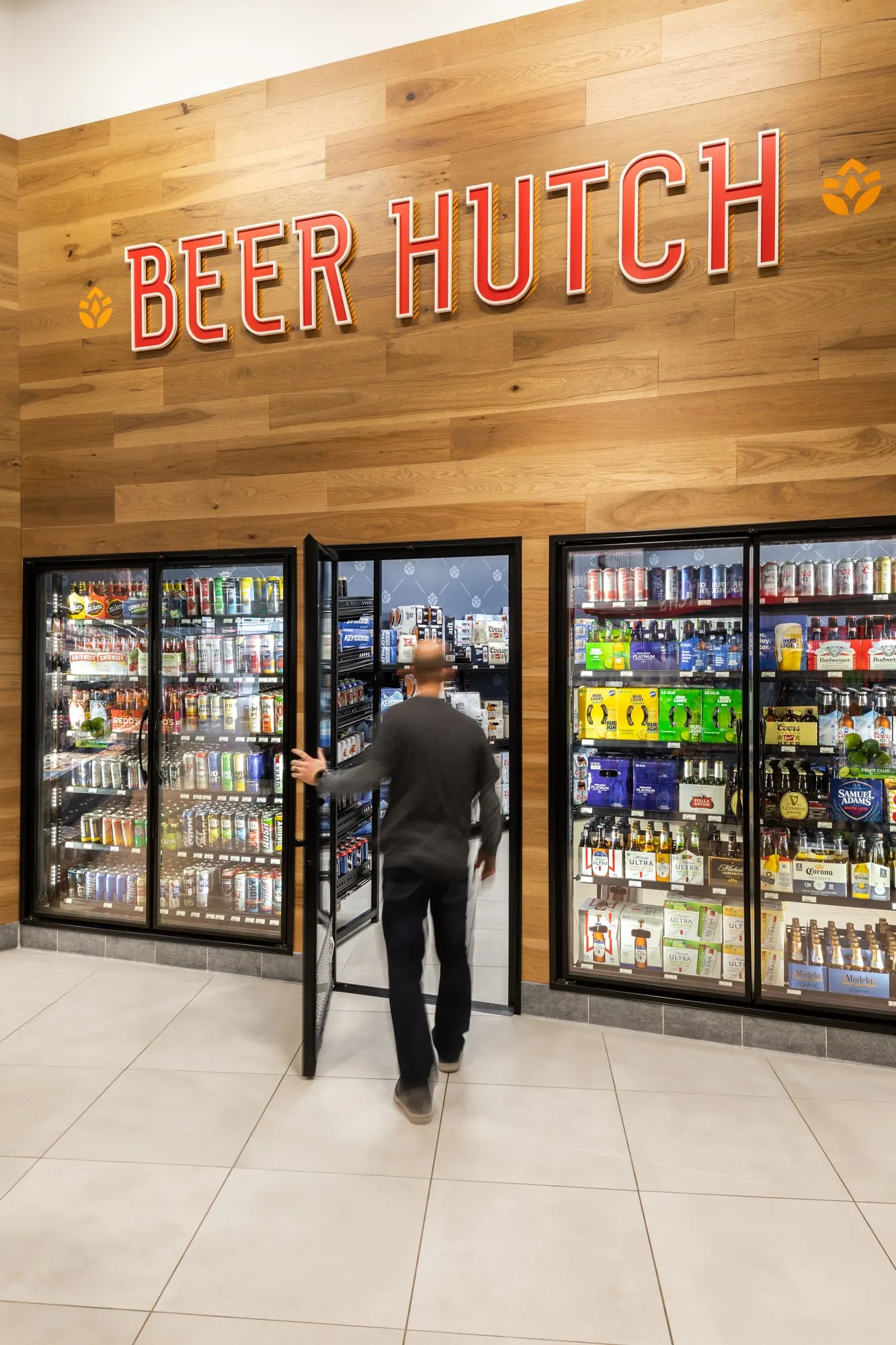 A photo of a person opening the door to a beer cave and walking in. The glass walls show stacks of beer packs kept inside. A sign over the door reads &quot;beer hutch.&quot;