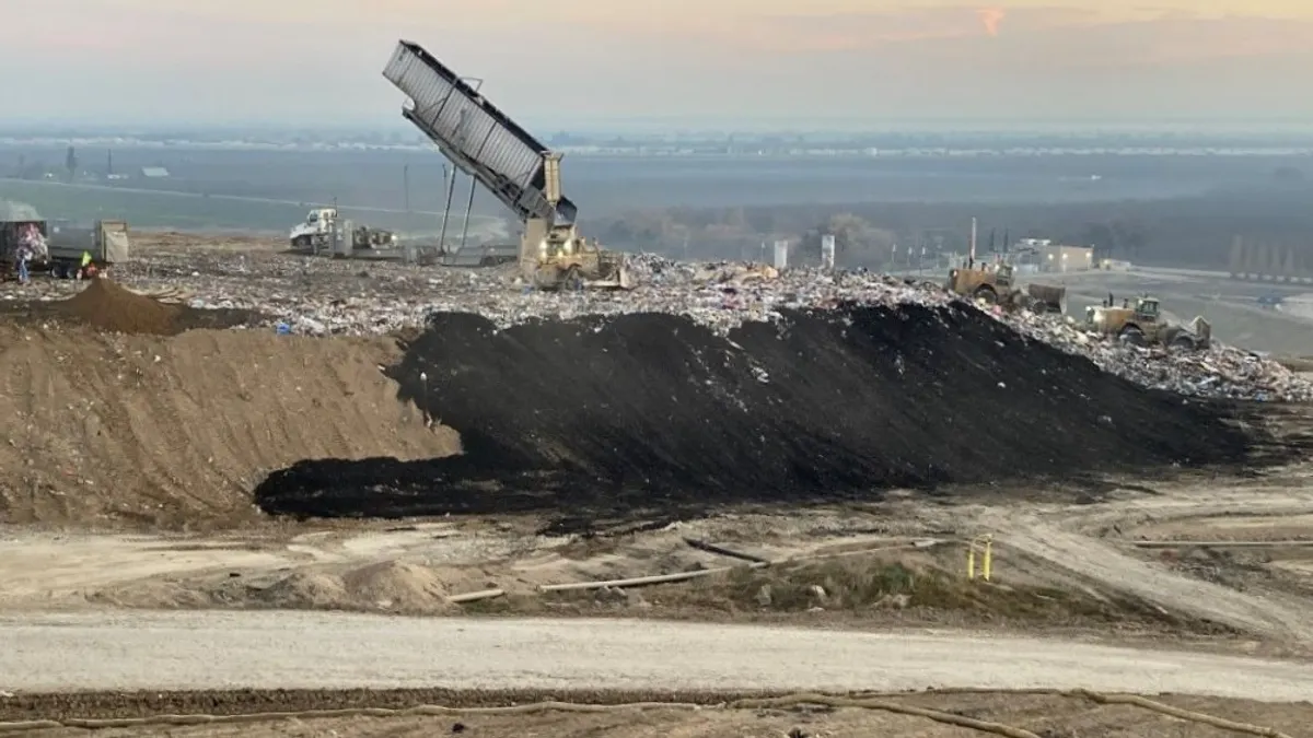 A truck dumps waste onto the open face of a landfill at dawn. Other trucks, workers, and landfill gas wells are visible.