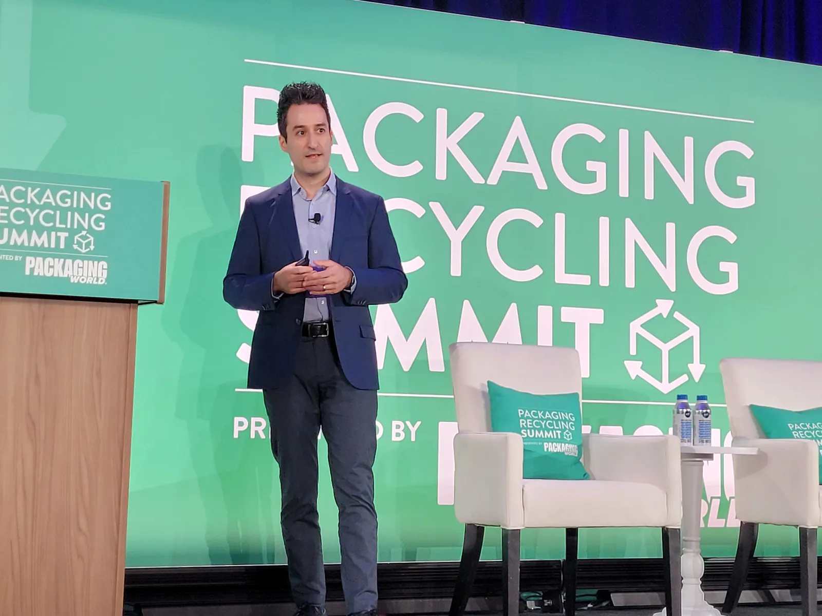 A person stands on a stage with a green screen behind them that says Packaging Recycling Summit.