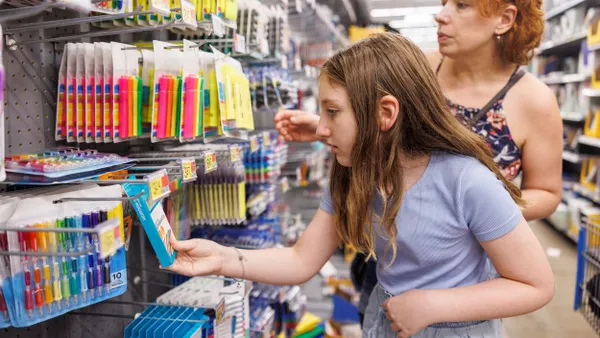 A parent and a child browse school supplies in a store aisle. The child is looking at a box of pens with the parent looking over their shoulder.