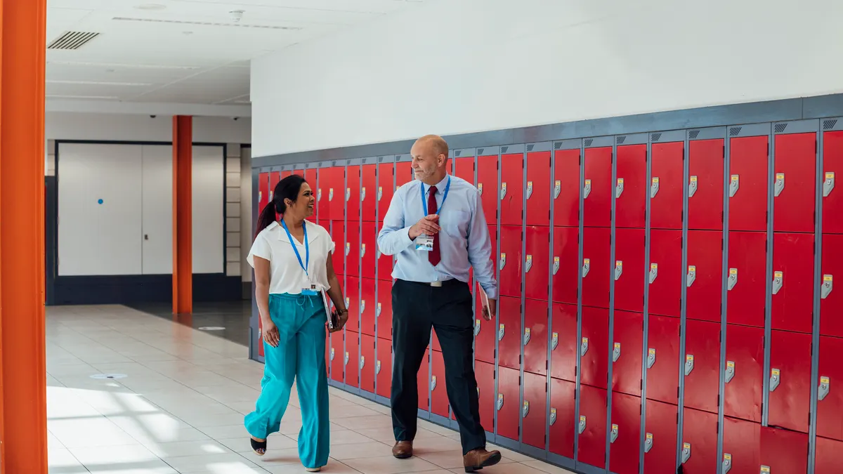 Two people wearing lanyards are walking down a school hallway lined with red lockers.