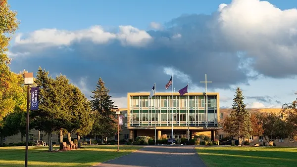 A campus building sits in the middle ground, behind a large cross and a flag pole with the U.S. flag.