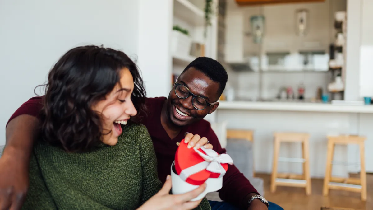 A boyfriend surprises his girlfriend with a Valentine's Day present, making her smile.