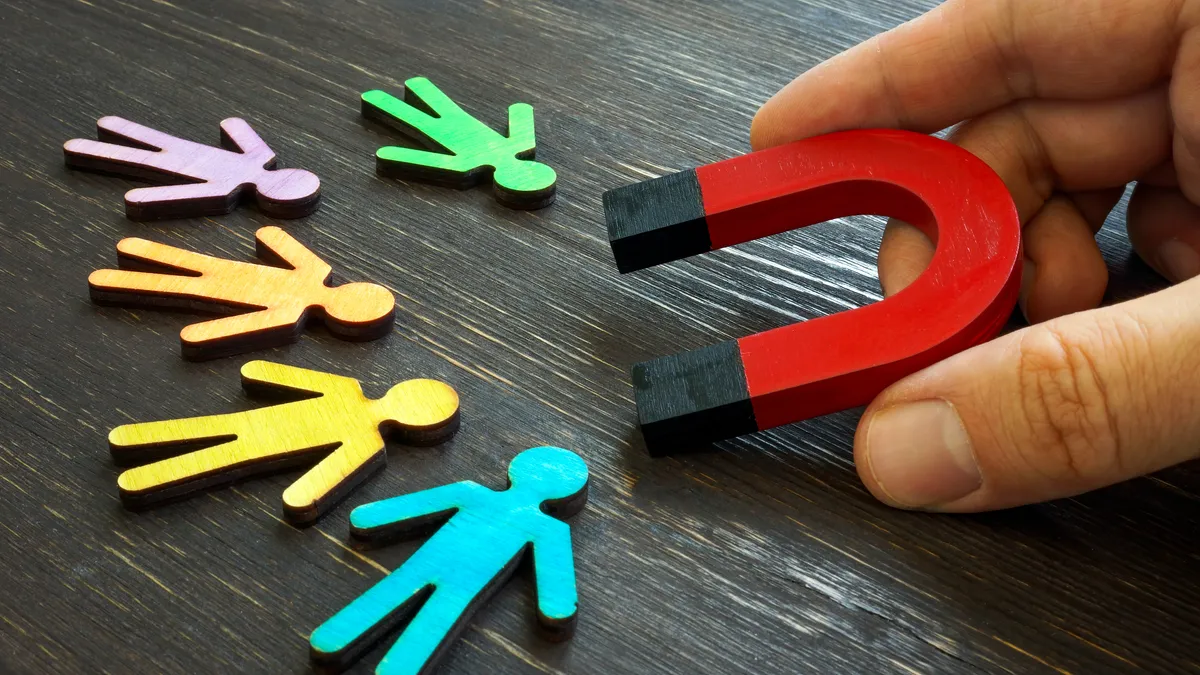 A person holds a magnet pointed at multi-colored wooden figures of people sitting on dark wood-grain table.