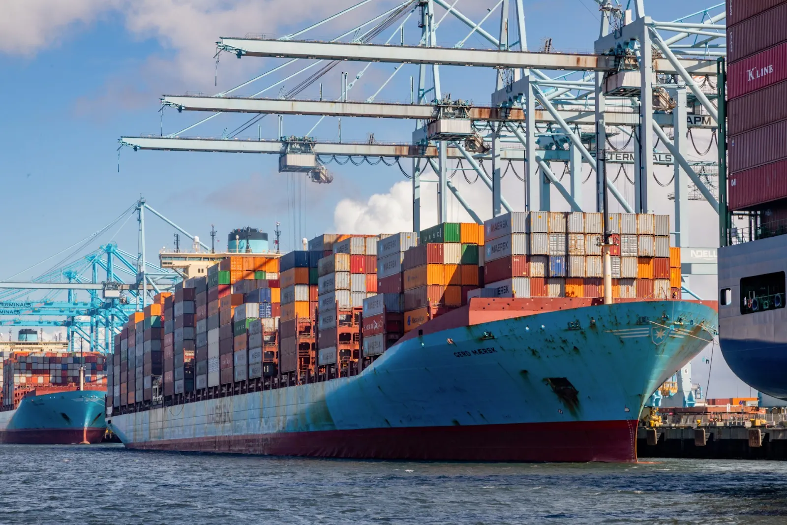 Dozens of multi-colored containers are stacked on a blue cargo ship docked at a port under cranes. The cargo ship is between two other vessels.