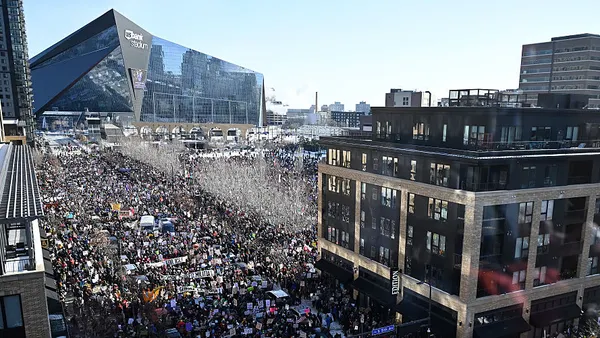 An aerial view depicts a downtown Minneapolis street packed with people protesting, with a football stadium in the background.