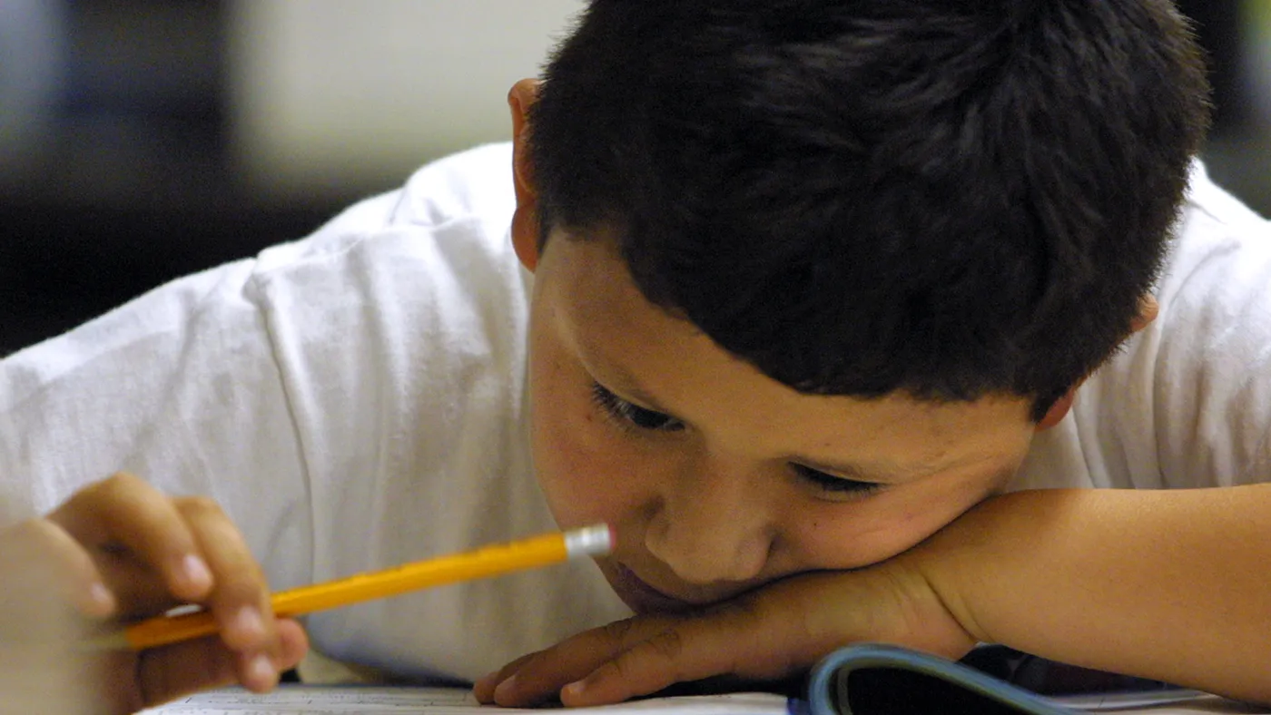 A young student holds a pencil while sitting at desk and looking down at a sheet of paper.