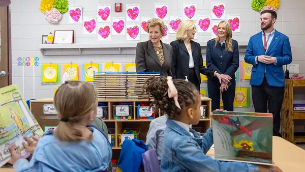 Four adults are standing in the back of a classroom. In the forefront are two students sitting at desks holding books open.