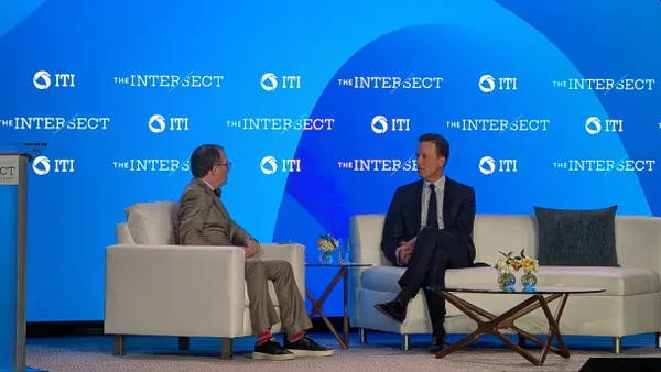 Two men sit on a stage in front of a blue backdrop bearing the logos of a conference and its sponsors