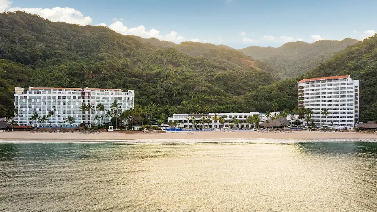 Three white resort buildings are seen along the beach with mountains behind them.