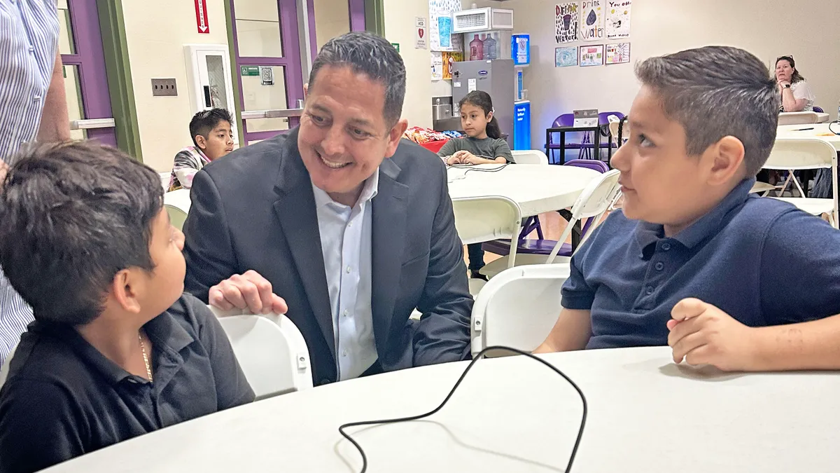 A Latino school leader in a suit kneels at a classroom table to speak with two boys.