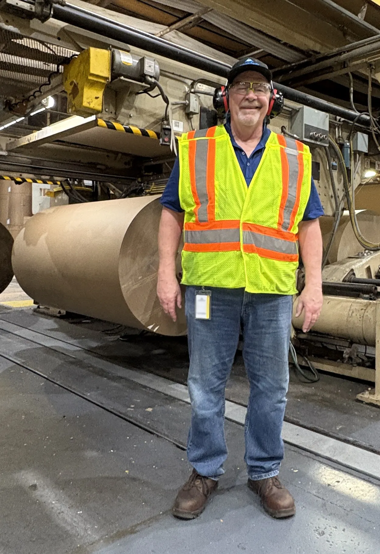 A person wearing safety goggles and a yellow vests stands in front of a large roll of paper in an industrial facility.