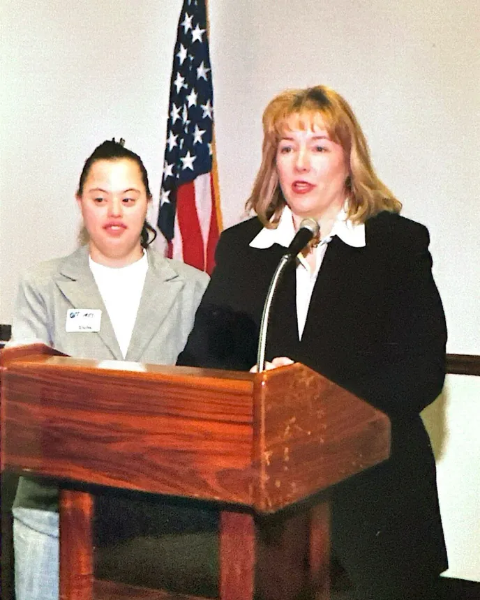 Two people stand inside a room behind a wooden podium. An American Flag on a pole is behind them.