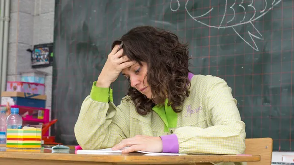 A person is sitting at a desk in a classroom. They look stressed. A chalkboard is behind them and there are papers on the desk.