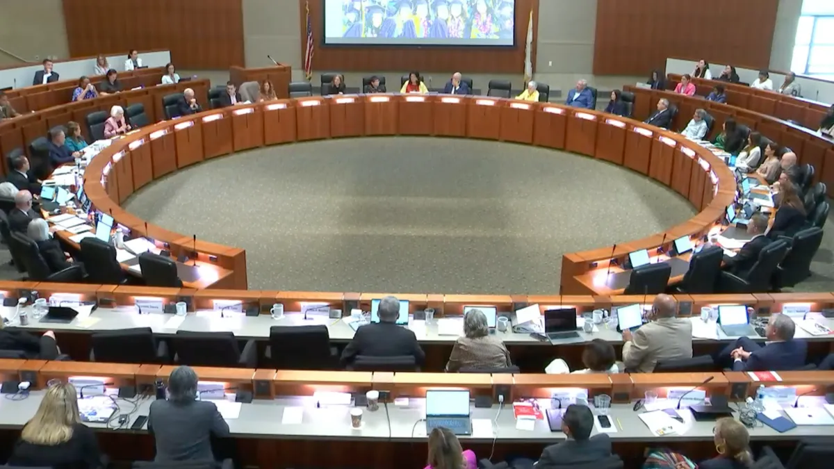 A ceiling-down view of a large meeting room with roughly a dozen people in desks facing one another.