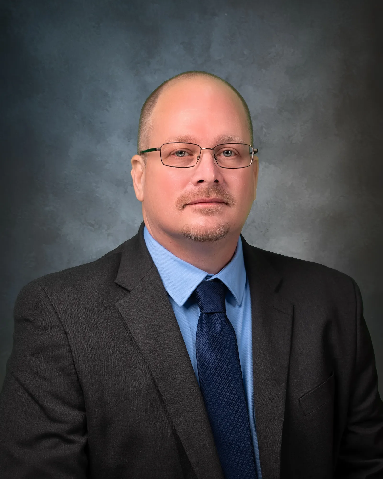 Headshot of man with wir rim glasses in a dark suit and blue shirt and tie.