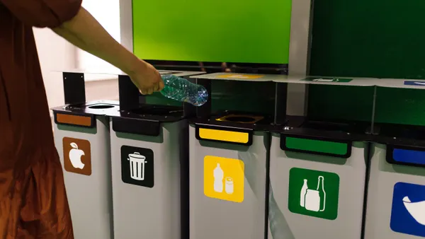 A person's arm is seen throwing a plastic bottle into an appropriate recycling bin, which is next to other waste bins.