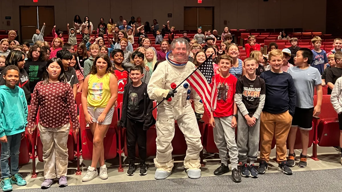 A man superintendent in an astronaut suit stands in an assembly hall packed with K-8 students.