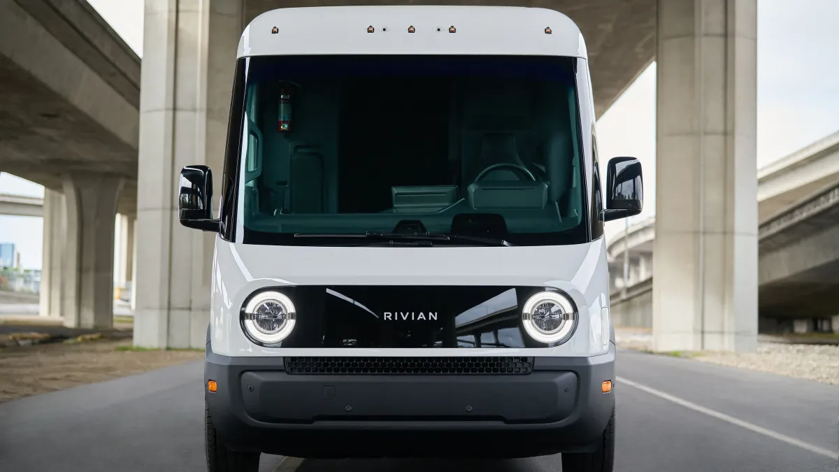 A front view showing the headlights and grill of s white Rivian's electric commercial delivery van parked under a highway overpass.