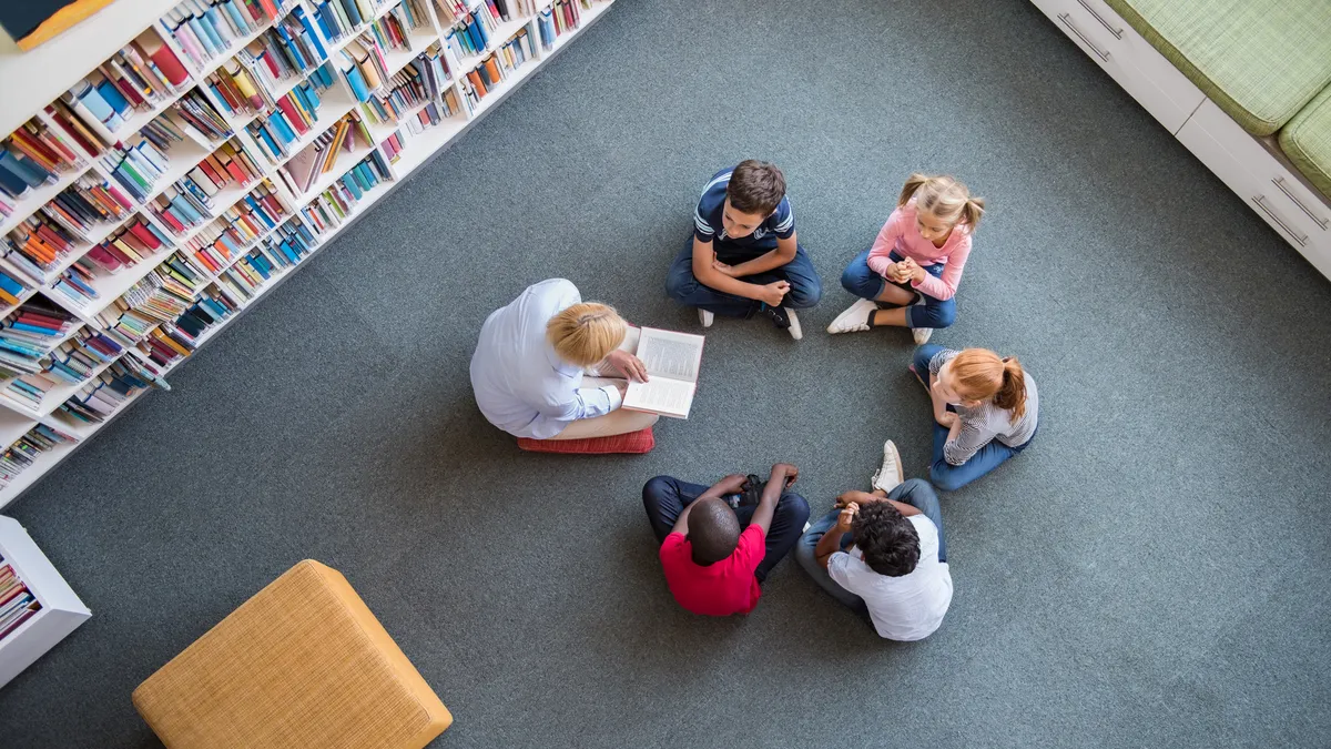 An above camera shot of an adult and a few students sitting on the floor in a circle in a library. The adult has a book open.