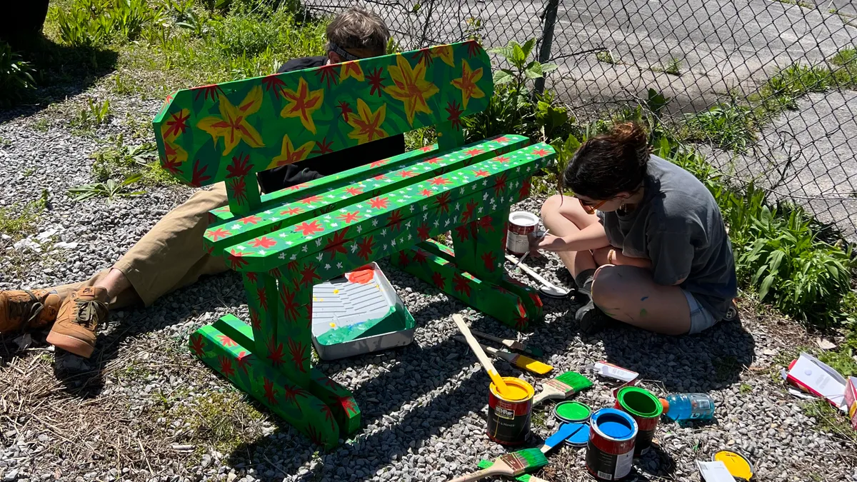 A person sits on the ground outside painting a colorful bench, while another lays behind the bench. Buckets of paint sit next to them.