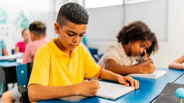 A young boy sits at a desk writing on a paper tablet. A young girl sits at a desk next to him, doing the same.