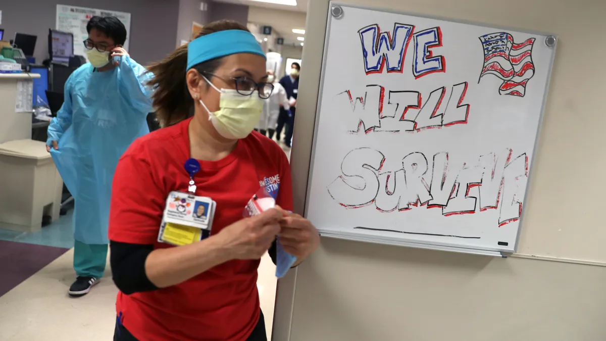 Nurses wearing masks walk through a hospital hall, in front of a "We Will Survive" sign.