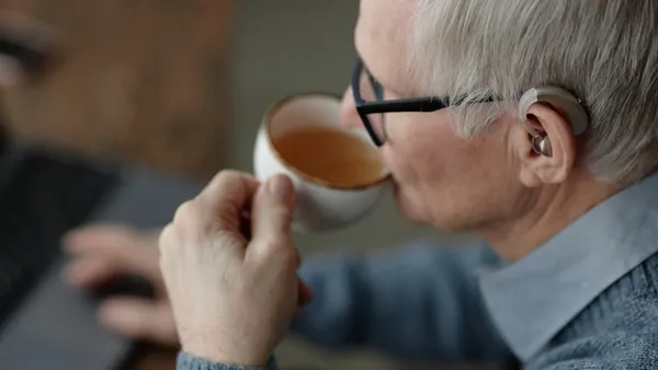 An older White man with a hearing aid sips tea.