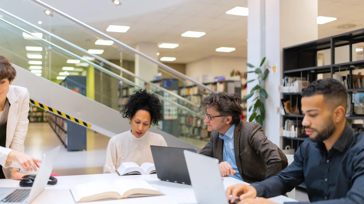 Adult students work and confer over laptops in a library