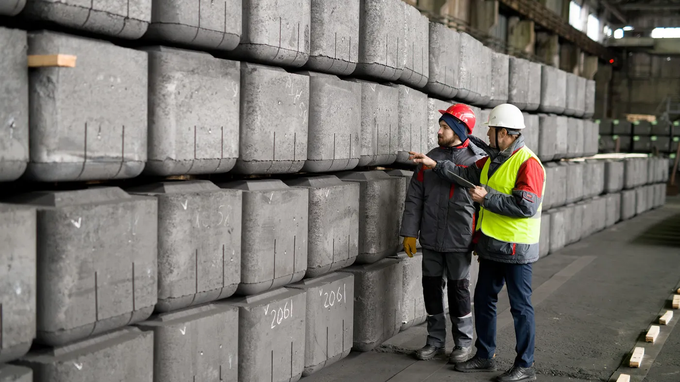 Two workers in safety gear conducting quality inspections of stacked large concrete squares inside an industrial plant.  