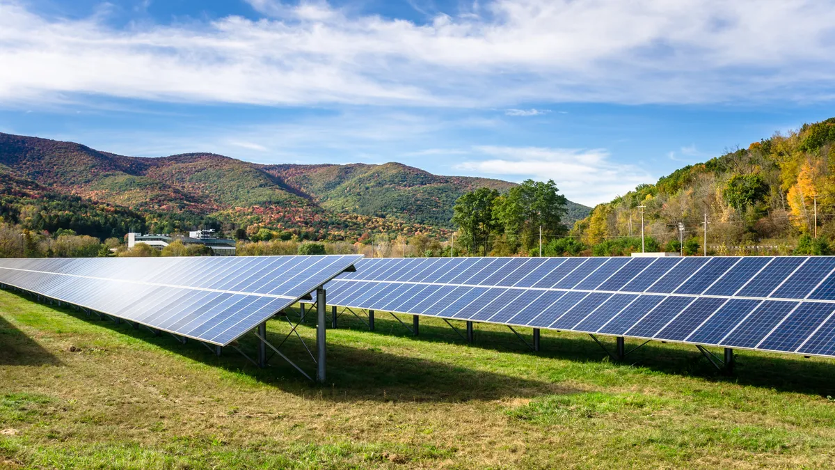 Solar Panels in a Grassy Field with Forested Hills in Background and Blue Sky with Clouds.