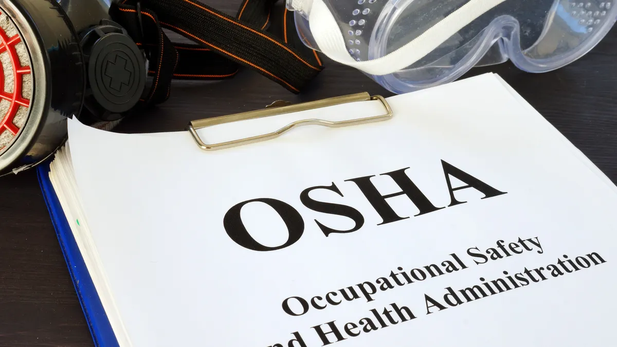 A blue clipboard with a white paper that says "OSHA, Occupational Safety and Health Administration," and protective gear lying above it.