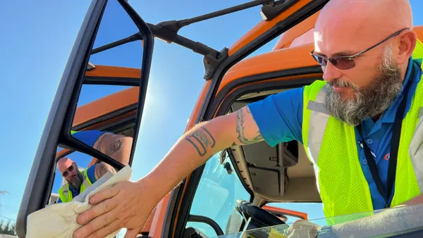 A person wearing a neon worker vest reaches out of the driver’s seat window of a large truck to wipe the truck’s side mirror. The person is reflected in the mirror.