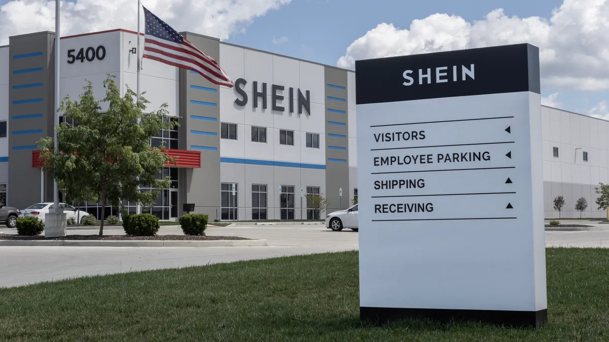 A closeup of a Shein sign directing people around its distribution center. The Shein building is seen in the background, with an American flag.