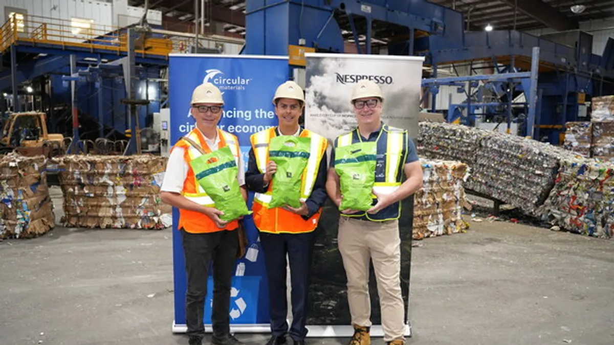 Three people in safety vests and hats hold green bags to recycle Nespresso coffee pods at an industrial facility.