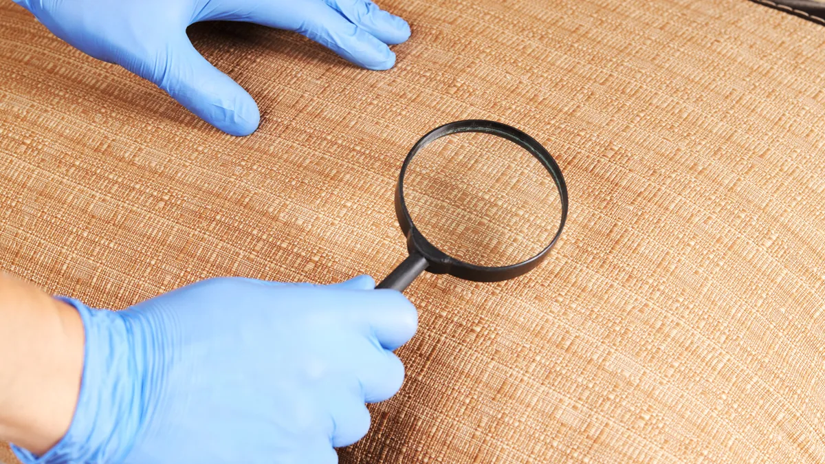 A person wears blue gloves and holds a magnifying glass over some upholstery.