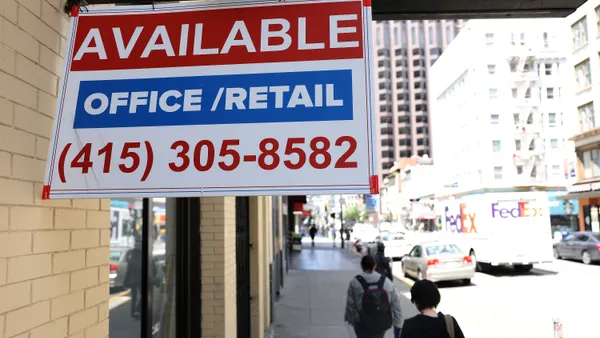 A sign outside of a city building reads "Available" and "office/retail" as people walk by on the sidewalk