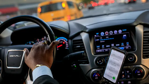 Car driver's hand on the wheel with a smartphone to the right.