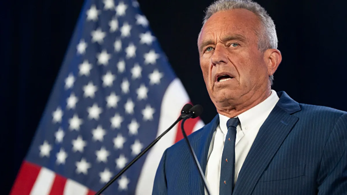 A person in a business suit speaks at a lectern with a U.S. flag in the background.