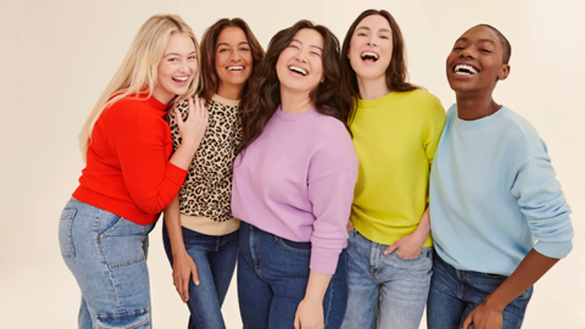 A smiling, diverse group of people, stand in a row, wearing colorful tops and blue jeans of various shades.