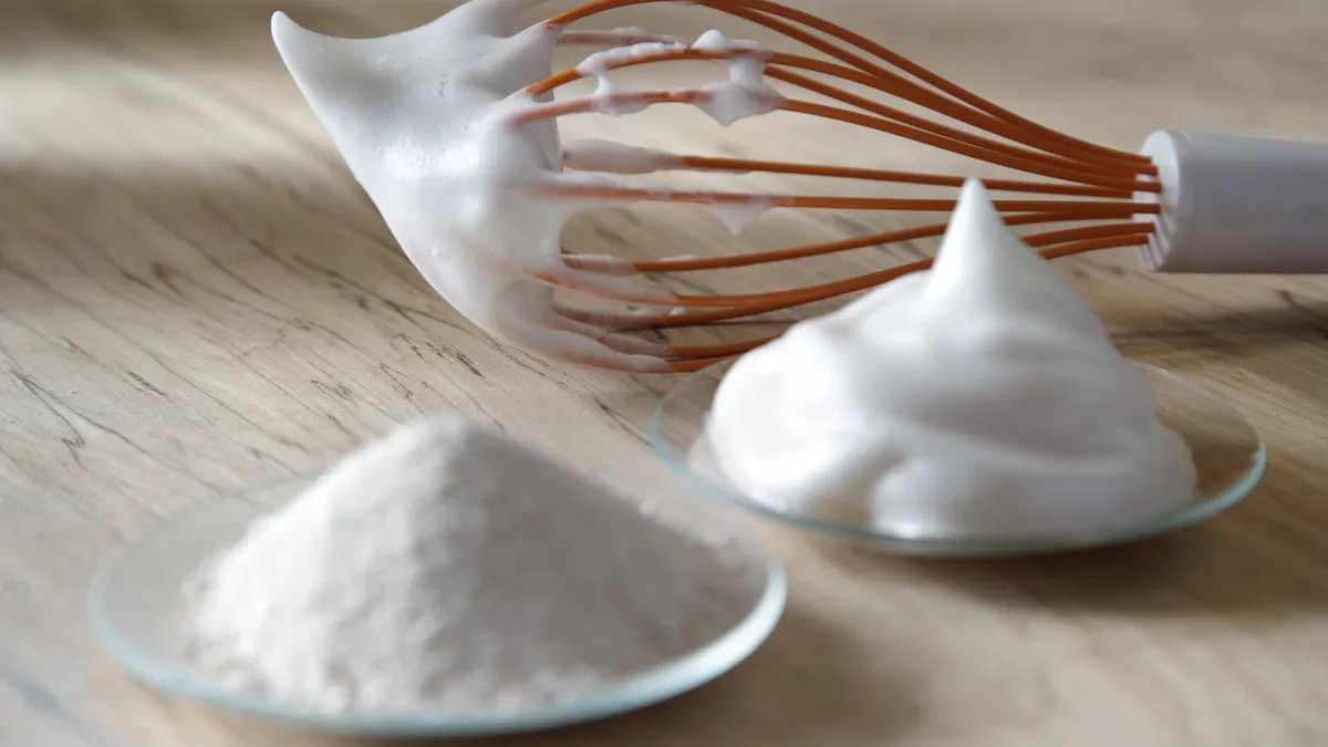 A dish of powdered egg white made through Onego Bio's precision fermentation sits on a counter, with a dish of whipped egg white and a whisk with whipped egg on it behind.
