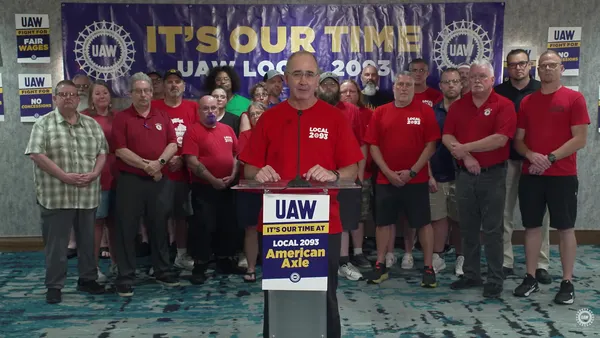 A person wearing a red shirt and glasses standing behind a podium, with people in red shirts standing in the background.