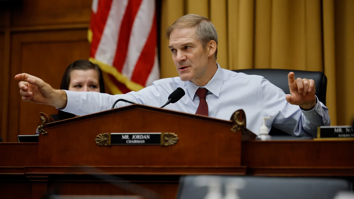 Jim Jordan sits behind a podium with both hands outstretched.