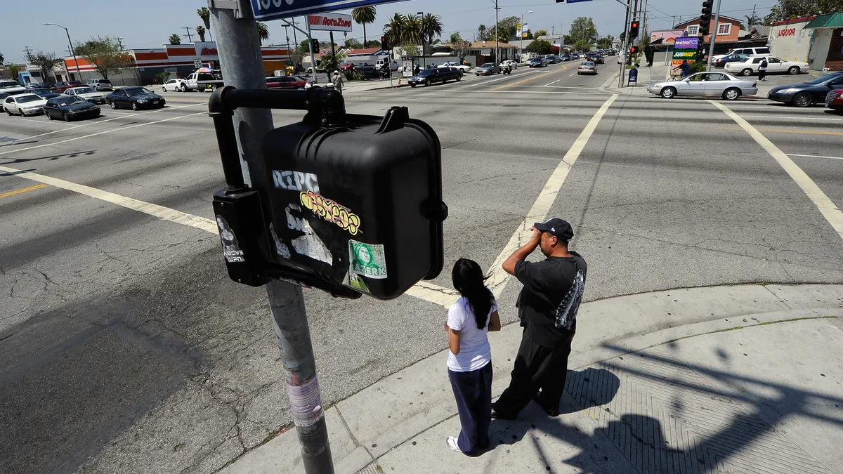 Two people wait on the curb before crossing a major intersection in Los Angeles.