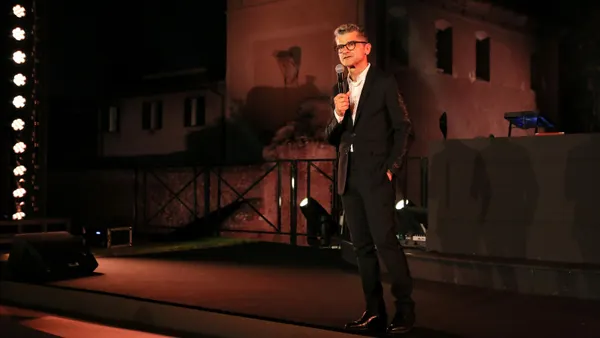 A man in a suit stands on a dimly lit stage holding a microphone.
